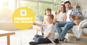 Familie sitzt lachend auf einem Sofa im Wohnzimmer junges Kind schaltet mit einer Fernbedienung den Fernseher um Hund sitzt auf dem Schoß des Mannes Text auf gelbem Kreis 18 PRODUKT DER WOCHE