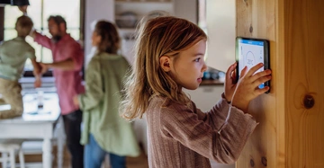 blondes Mädchen in graulila Strickpulli bedient das Touchpanel an der Wohnzimmerwand. Im Hintergrund Mama, Papa und Bruder beim Herumtollen.