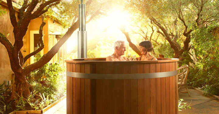 Galerie-Bild 6: Zwei Personen entspannen in einem Holzbadefass und halten Getränke. Das Fass steht in einem sonnigen Garten mit vielen Bäumen und Pflanzen, und ein Haus ist im Hintergrund sichtbar.