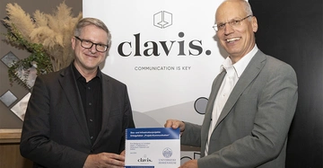 Frank Brettschneider von der Uni Stuttgart und clavis-Geschäftsführer Ulrich Müller halten lächelnd ihre Studienarbeit in die Kamera, im Hintergrund ein Aufsteller mit dem Schriftzug "clavis"