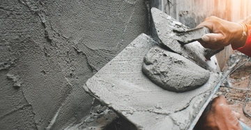 Mann mit Maurerkelle und frischem Mörtel beim Verputzen einer Außenwand. Großaufnahme, alles Grau in Grau.