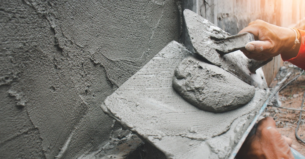 Mann mit Maurerkelle und frischem Mörtel beim Verputzen einer Außenwand. Großaufnahme, alles Grau in Grau.