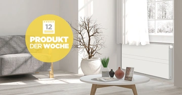 Graues Sofa steht neben einem Heizkörper in hellem Raum mit Fenster und kahlem Baum; niedriger Tisch mit Dekorationsobjekten steht zentral. Text auf gelbem Kreis: 12 PRODUKT DER WOCHE.