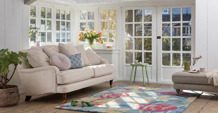 Beiges Sofa mit Dekokissen steht in einem hellen Wohnzimmer neben einem runden kleinen Tisch vor großen Sprossenfenstern, einem blumigen Teppich und einem Hocker unter einer Blumenvase.