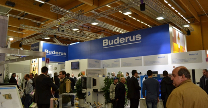 Galerie-Bild 23: Mehrere Personen stehen und unterhalten sich in einer Ausstellungshalle vor einem blauen Stand der Firma Buderus der verschiedene Exponate und Schautafeln zeigt