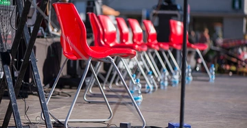 Mehrere rote Plastikstühle stehen in einer Reihe auf einer Bühne Wasserflaschen sind auf dem Boden verteilt im Hintergrund sind Unschärfe und verschiedene Gegenstände zu sehen
