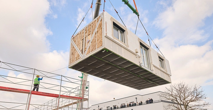 Modul mit Tür und zwei Fenstern wird mit Kran verhoben, Arbeiter auf Gerüst gibt Anweisungen, im Hintergrund zahlreiche Zuschauer auf Balkon eines Gebäudes