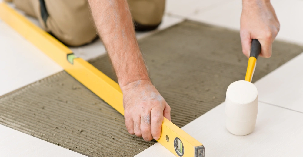 Hande richten mit Wasserwaage und Gummihammer Fliesen auf feuchtem Zement aus Knieende Person im Hintergrund helle Umgebung