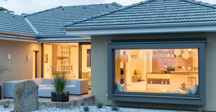 Modernes Einfamilienhaus zeigt hell erleuchteten Innenraum durch großes Fenster während draußen eine Steinterrasse mit Kieselsteinen und Pflanzen sichtbar ist das Haus hat ein Walmdach mit dunkelgrauen Ziegeln