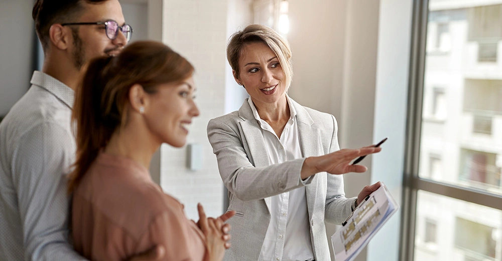 Immobilienmaklerin zeigt jungem Paar attraktive Wohnung mit großen Fenstern und hellem Ausblick. Professionelle Immobilienberatung beim Besichtigungstermin.