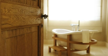 Holztür geöffnet Badezimmer freistehende Badewanne stehend ein Handtuch am Rand hängend Sonnenlicht durch ein großes Fenster einfallend