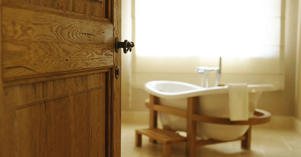 Holztür geöffnet Badezimmer freistehende Badewanne stehend ein Handtuch am Rand hängend Sonnenlicht durch ein großes Fenster einfallend