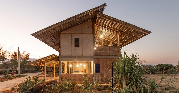 Haus aus Holz und Bambus mit großer Terrasse unter dem Dach und hell erleuchteten Fensterfronten. Umgebung Schilf, Palmen, im Hintergrund das Meer. 