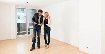Ein Mann in Jeans und Sakko und eine Frau in Jeans und hellem Sakko stehen in einem leeren großen Raum und schauen sich Pläne an, während sie etwas besprechen.