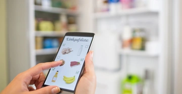 Eine Hand hält ein Smartphone zeigt eine Einkaufsliste mit Bildern in der Küche vor dem offenen Kühlschrank. Der Bildschirmtext lautet Einkaufsliste. Markierte Artikel: Huhn Milch Schinken Bananen.