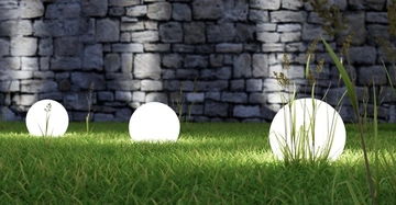 Leuchtkugeln liegen auf Gras und leuchten, während sie von großen Grasbüscheln umgeben sind. Im Hintergrund ist eine grobe Steinmauer zu sehen, die die Szenerie ergänzt.