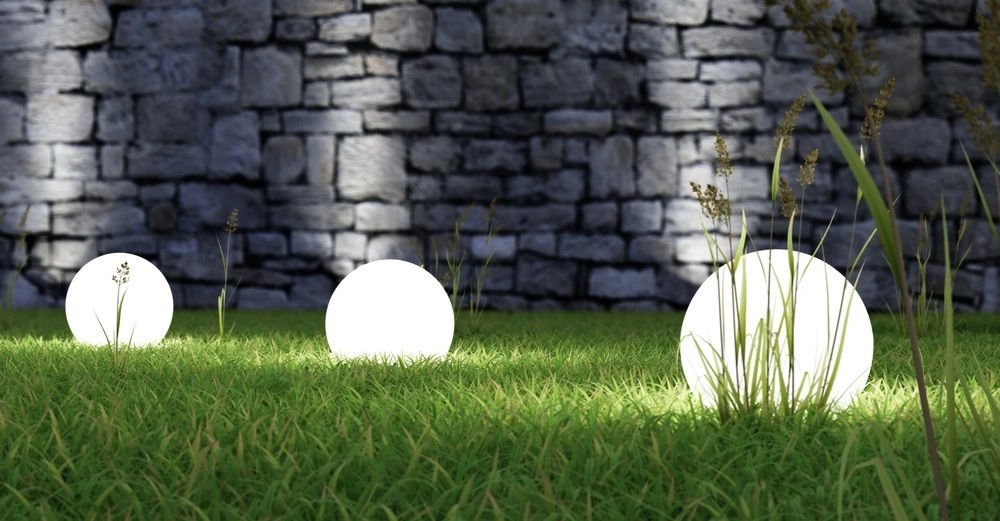 Leuchtkugeln liegen auf Gras und leuchten, während sie von großen Grasbüscheln umgeben sind. Im Hintergrund ist eine grobe Steinmauer zu sehen, die die Szenerie ergänzt.
