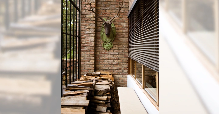 Galerie-Bild 4: Ein Hirschkopf-Trophäe hängt an einer Backsteinwand über gestapeltem Brennholz in einem engen Raum mit Fenstern zu einer Seite und einem kleinen Sitzbank davor.