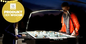 Zwei Personen spielen Tischfußball bei Nacht mit Beleuchtung. Großes gelbes Symbol mit Text 15 PRODUKT DER WOCHE im Vordergrund links.