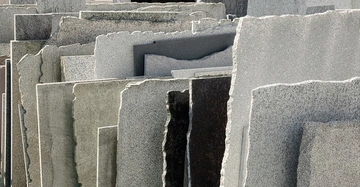 Mehrere große steinerne Platten stehen aufrecht und dicht nebeneinander gelagert an einem Freiluftlagerplatz. Die Platten sind unterschiedlich gefärbt und haben teilweise raue, unebene Kanten.