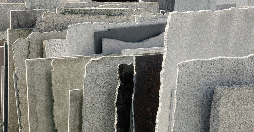 Mehrere große steinerne Platten stehen aufrecht und dicht nebeneinander gelagert an einem Freiluftlagerplatz. Die Platten sind unterschiedlich gefärbt und haben teilweise raue, unebene Kanten.