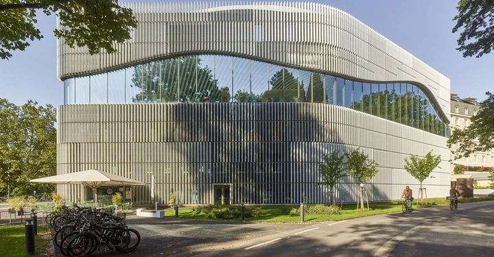 Ein modernes Gebäude mit geschwungener Fassade steht im Freien von Bäumen umgeben Radfahrer fahren auf einer Straße im Vordergrund Fahrräder sind in einem Ständer abgestellt Ein Café im Erdgeschoss