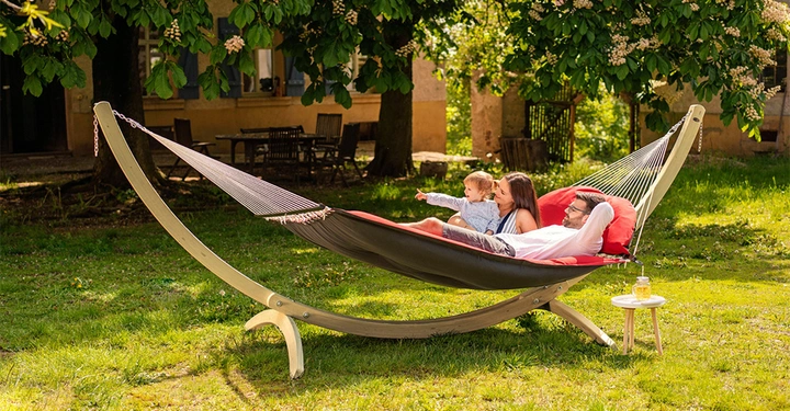 Galerie-Bild 3: Eine Familie liegt entspannt auf einer Hängematte und spielt mit ihrem Kind im Garten mit grüner Rasenfläche und Bäumen im Hintergrund. Ein Beistelltisch mit Getränk steht daneben.