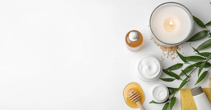 Kerzenlicht brennt neben kosmetischen Produkten und Honig auf Honiglöffel, umgeben von grünen Blättern, auf einer weißen Oberfläche.