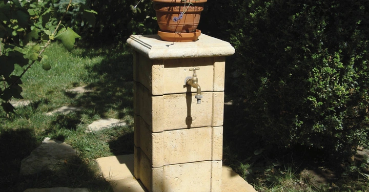 Galerie-Bild 4: Ein Blumentopf mit Lavendel steht auf einem steinernen Sockel mit Wasserhahn im grünen Garten neben Büschen und Bäumen auf dem Gras mit Steinplatten.