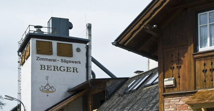 Galerie-Bild 9: Ein weißes Gebäude mit der Aufschrift Zimmerei Sägewerk BERGER zeigt Schornsteine und Metallstrukturen im Freien neben einem älteren, hölzernen Gebäude mit Fenstern und Dachgauben.