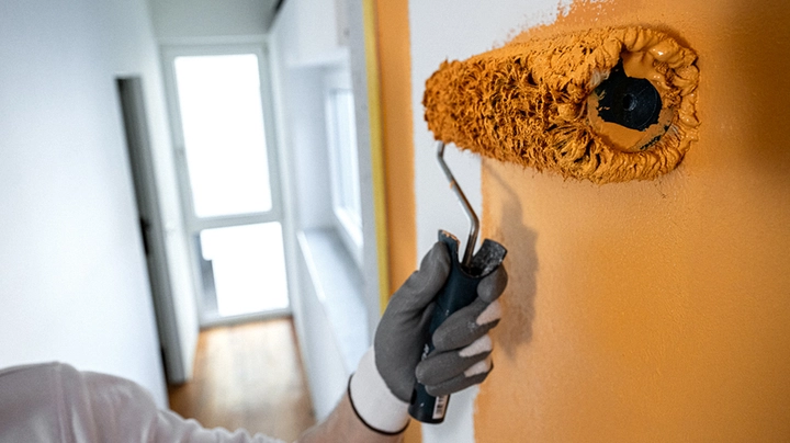 Galerie-Bild 2: Nahaufnahme einer Hand mit Arbeitshandschuh, die mit einer Farbrolle eine Wand in einem warmen Orange- oder Ockerton streicht. Im Hintergrund ist ein heller, schmaler Flur mit Holzboden und einer Tür am Ende zu sehen.