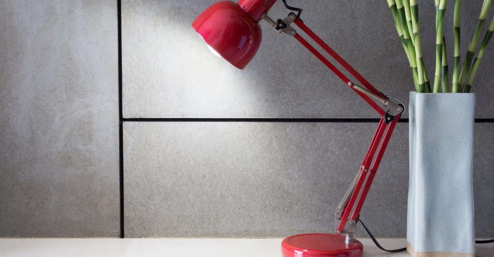 Rote Schreibtischlampe beleuchtet Tischoberfläche neben hoher Vase mit Bambus im modernen grauen Raum.