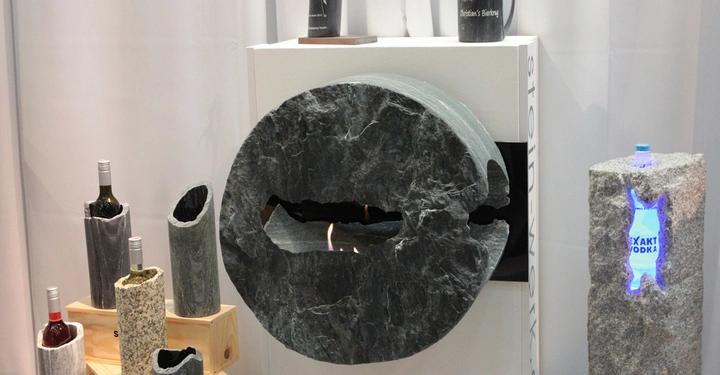 Galerie-Bild 4: Großer grauer Steinring mit integrierter Flamme hängt vor weißem Hintergrund Weinflaschen werden in zylinderförmigen Steinen auf Holzstufen angezeigt beleuchtete Wodkaflasche in vertikalem Steinzylinder platziert Text auf dem Bild: Christian's Bierkrug STEIN-W EXAKT WODKA