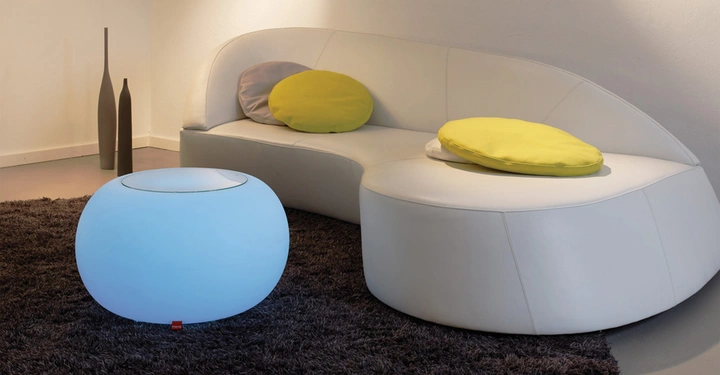 Heller Couchtisch leuchtet blau und steht auf einem dunklen Teppich; daneben ein geschwungenes weißes Sofa mit gelben Kissen in einem minimalistischen Wohnzimmer.