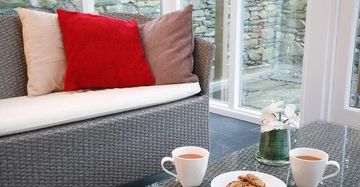 Ein graues Sofa mit bunten Kissen steht in einem Raum mit großen Fenstern. Ein Glastisch davor trägt zwei Tassen Tee, einen Teller Kekse und eine Vase mit Blumen.