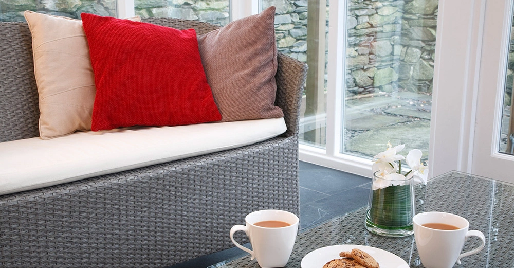 Ein graues Sofa mit bunten Kissen steht in einem Raum mit großen Fenstern. Ein Glastisch davor trägt zwei Tassen Tee, einen Teller Kekse und eine Vase mit Blumen.