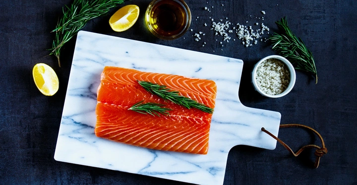 Ein Stück roher Lachs liegt auf einem Marmorschneidebrett mit Rosmarinzweigen darauf. Drumherum liegen Zitronenspalten, Rosmarinzweige, eine Schale Olivenöl und eine Schale grobes Salz auf einem dunklen Untergrund.