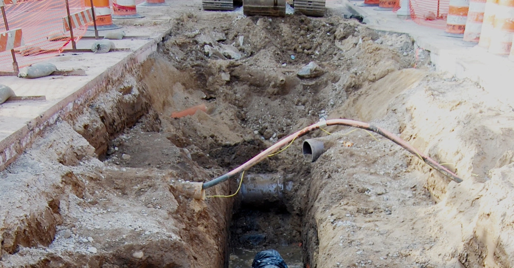 Ein Bagger gräbt einen tiefen Graben in einer Baustelle aus Straßenarbeiten mit Verkehrshütchen und Maschendrahtzäunen im Hintergrund. Rohre und Kies im Graben.