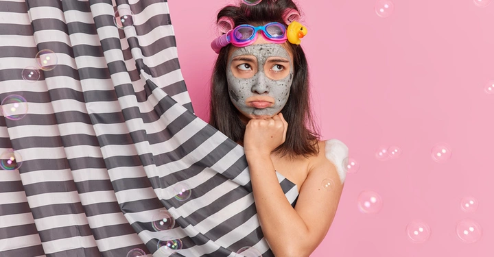 junge Frau mit braunem Haar, Lockenwicklern und einer Gesichtsmaske steht nachdenklich halb verdeckt hinter grau-weiß gestreifetem Duschvorhang, Hintergrund rosarot