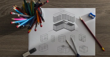 Zeichnung einer Küche auf Papier mit Bleistiften, Zirkel und Spitzer auf einem Holztisch. Links ein Bleistifthalter mit bunten Stiften.