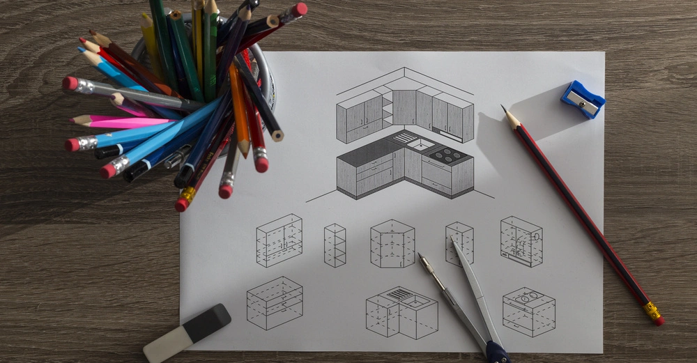 Zeichnung einer Küche auf Papier mit Bleistiften, Zirkel und Spitzer auf einem Holztisch. Links ein Bleistifthalter mit bunten Stiften.
