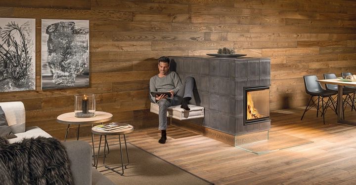 Galerie-Bild 1: Ein Mann sitzt lesend auf einem Podest vor einem modernen Kamin in einem Wohnzimmer mit Holzwänden auf dem Tisch eine Kerze im Hintergrund ein Esstisch mit Stühlen.