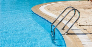 Metall-Leiter taucht teilweise ins leuchtend blaue Wasser eines Schwimmbeckens ein Amphitheaterförmige Terrassierung mit Braunstufen umgibt den Pool