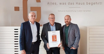 Drei Männer stehen vor einer Wand der mittlere Mann hält eine Auszeichnung in den Händen Hintergrund zeigt Schriftzug Alles was das Haus begehrt Exklusive Ausstattung in allen Varianten