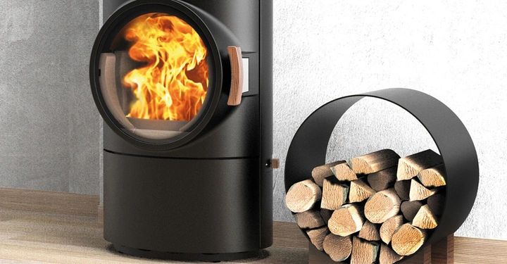 Ein schwarzer Kaminofen brennt und erzeugt Flammen Holzhaufen in einem runden Ständer steht daneben in einem Zimmer mit Holzboden und grauer Wand.