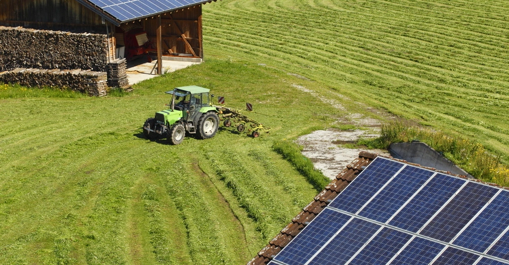 Ein Traktor mäht das Gras auf einem großen grünen Feld gelegen bei einer Scheune mit gestapeltem Holz und Solarpaneelen auf dem Dach im ländlichen Gebiet