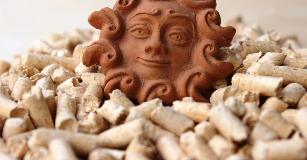 Ein braunes, lächelndes Sonnengesicht aus Ton liegt auf einem Haufen heller Holzpellets in einem unscharfen, neutralen Hintergrund.