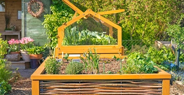 Hochbeet aus hellem Holz mit aufgeklapptem Dach, Schuppen im Hintergrund, schöner Garten, viel Obst und Gemüse im Hochbeet.