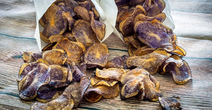 Lila Kartoffelchips fallen aus weißen Papiertüten und liegen verstreut auf einer Holzoberfläche
