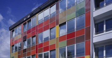 Ein mehrstöckiges Gebäude zeigt eine Fassade mit farbenfrohen, quadratischen Paneelen aus Glas oder Kunststoff. Das Gebäude steht unter blauem Himmel mit einigen weißen Wolken.